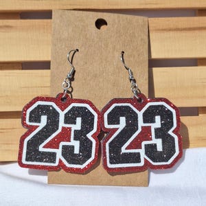 May include: A pair of dangle earrings featuring the number 23 in black glitter with a white outline, set against a red glitter background. The earrings are attached to a brown card and include silver-toned metal hooks for pierced ears.