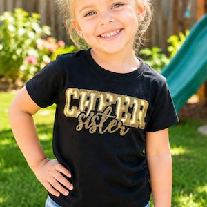 K&ouml;nnte beinhalten: Ein schwarzes Kurzarm-T-Shirt mit dem Wort CHEER in gro&szlig;en, metallisch goldenen Blockbuchstaben mit glitzernder Textur, platziert &uuml;ber dem Wort sister, das in einer kleineren, kursiven, glitzernden goldenen Schrift auf der Brust steht.