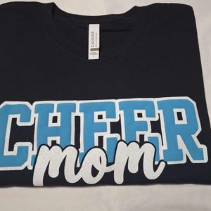 K&ouml;nnte beinhalten: Ein schwarzes Kurzarm-T-Shirt mit dem Wort CHEER in gro&szlig;en, hellblauen Blockbuchstaben mit wei&szlig;er Umrandung und dem Wort mom in wei&szlig;er Schreibschrift darunter. Das T-Shirt hat ein wei&szlig;es Nackenetikett mit dem Markennamen CANVAS.