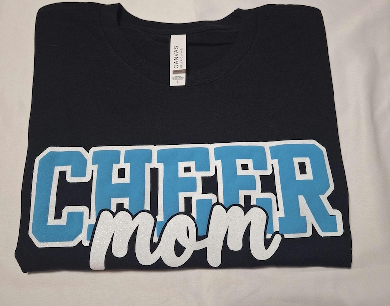 K&ouml;nnte beinhalten: Ein schwarzes Kurzarm-T-Shirt mit dem Wort CHEER in gro&szlig;en, hellblauen Blockbuchstaben mit wei&szlig;er Umrandung und dem Wort mom in wei&szlig;er Schreibschrift darunter. Das T-Shirt hat ein wei&szlig;es Nackenetikett mit dem Markennamen CANVAS.