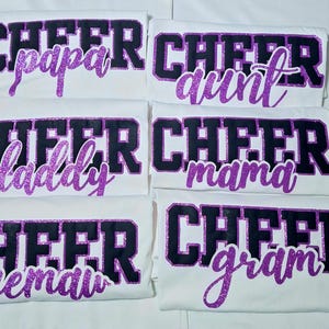 K&ouml;nnte beinhalten: Sechs wei&szlig;e T-Shirts mit dem Wort CHEER in gro&szlig;en schwarzen Blockbuchstaben mit violetten Glitzerumrandungen. Unter dem Wort CHEER zeigt jedes T-Shirt ein anderes Wort in violetter Glitzerschrift: papa, aunt, daddy, mama, memaw und gram.