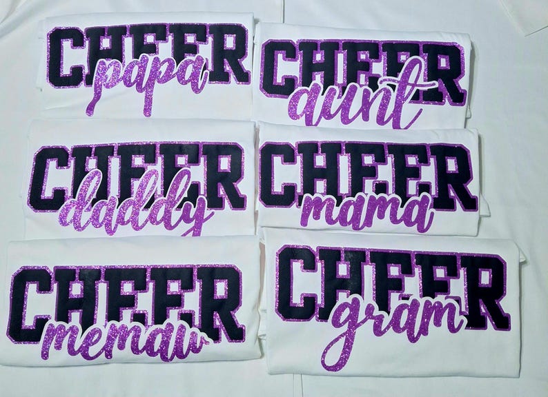 K&ouml;nnte beinhalten: Sechs wei&szlig;e T-Shirts mit dem Wort CHEER in gro&szlig;en schwarzen Blockbuchstaben mit violetten Glitzerumrandungen. Unter dem Wort CHEER zeigt jedes T-Shirt ein anderes Wort in violetter Glitzerschrift: papa, aunt, daddy, mama, memaw und gram.