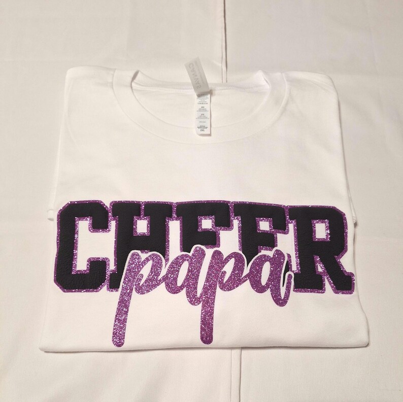 K&ouml;nnte beinhalten: Ein wei&szlig;es T-Shirt mit kurzen &Auml;rmeln, das das Wort CHEER in gro&szlig;en schwarzen Blockbuchstaben mit violettem Glitzerumriss zeigt, sowie das Wort papa in violetter Glitzerschrift, das &uuml;ber dem unteren Teil des schwarzen Textes liegt.