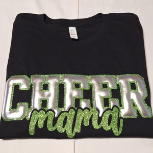 K&ouml;nnte beinhalten: Ein schwarzes Kurzarm-T-Shirt mit dem Wort CHEER in gro&szlig;en silbernen Blockbuchstaben mit einem gr&uuml;nen Glitzer-Umriss und dem Wort mama in gr&uuml;ner Glitzer-Schreibschrift, das direkt unter dem Wort CHEER platziert ist.