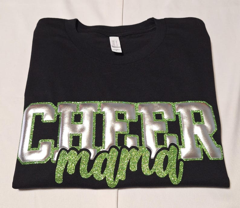 K&ouml;nnte beinhalten: Ein schwarzes Kurzarm-T-Shirt mit dem Wort CHEER in gro&szlig;en silbernen Blockbuchstaben mit einem gr&uuml;nen Glitzer-Umriss und dem Wort mama in gr&uuml;ner Glitzer-Schreibschrift, das direkt unter dem Wort CHEER platziert ist.