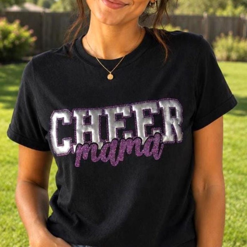 K&ouml;nnte beinhalten: Ein schwarzes T-Shirt mit kurzen &Auml;rmeln, das das Wort CHEER in gro&szlig;en, metallisch silbernen Blockbuchstaben mit violettem Glitzerumriss zeigt, sowie das Wort mama in violetter Glitzerschrift direkt unter dem silbernen Text.