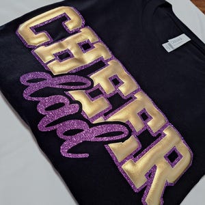 K&ouml;nnte beinhalten: Ein schwarzes T-Shirt mit dem Text CHEER DAD. Das Wort CHEER besteht aus gro&szlig;en, metallisch goldenen Blockbuchstaben mit einem violetten Glitzerumriss. Das Wort DAD ist in einer violetten Glitzerschrift &uuml;ber die goldenen Buchstaben gelegt.