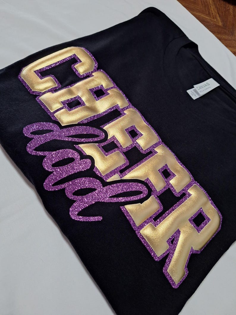 K&ouml;nnte beinhalten: Ein schwarzes T-Shirt mit dem Text CHEER DAD. Das Wort CHEER besteht aus gro&szlig;en, metallisch goldenen Blockbuchstaben mit einem violetten Glitzerumriss. Das Wort DAD ist in einer violetten Glitzerschrift &uuml;ber die goldenen Buchstaben gelegt.