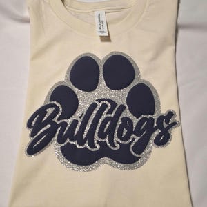 May include: A cream-colored t-shirt featuring a dark navy blue paw print graphic with a silver glitter outline. The word Bulldogs is written in dark navy blue script across the center of the paw print. A Bella+Canvas brand tag is visible at the neck.