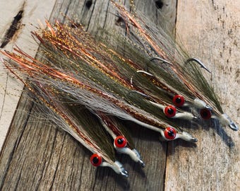 May include: A collection of five fishing lures featuring long, flowing olive green and white synthetic fibers mixed with shimmering copper tinsel. Each lure is equipped with a silver metal hook and a prominent, round, bright red eye with a black pupil.