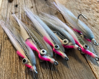 May include: Six fishing flies with gray, white, and pink accents. Each fly has a silver hook and a painted pink head with a large eye. The flies are arranged on a weathered wooden surface.