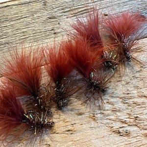 May include: Close-up of six handmade fishing flies. Each fly features a dark brown hook, a body of brown and iridescent material, and a large, reddish-brown feather plume. The flies are arranged on a weathered wooden surface.