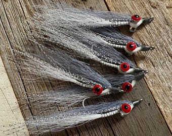 May include: Five fishing flies with gray and white synthetic fibers and red eyes are arranged on a weathered wooden surface. The flies have a streamlined design, with a silver thread accenting the body. The background is a rustic wooden plank.