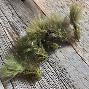 May include: Close-up of several olive green fishing flies with feathery bodies and silver hooks. The flies are arranged on a weathered wooden surface, with natural light casting shadows. The flies are designed for fly fishing.