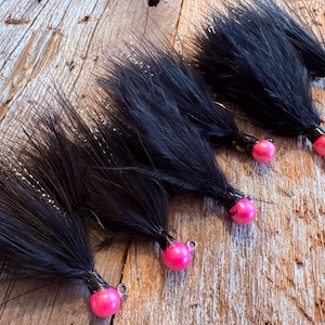 May include: Several black fishing lures with pink, round heads are displayed. The lures have a fluffy, feather-like appearance and are arranged on a weathered, wooden surface. The pink heads have a small metal loop.
