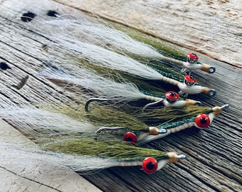 May include: Close-up of five fishing flies. Each fly features a white and olive green feathered body, a red eye, and a silver hook. The flies are arranged on a weathered wooden surface, showcasing their intricate details and craftsmanship.