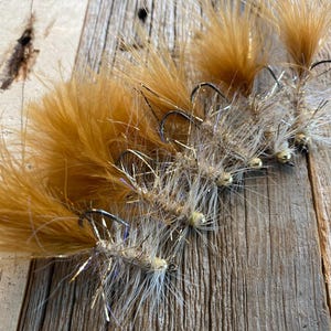 May include: Close-up of several handmade fishing flies. The flies feature brown and tan feathers, silver tinsel, and black hooks. They are arranged on a weathered wooden surface, showcasing the intricate details of each fly.