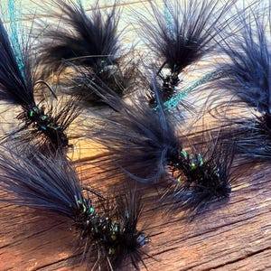 May include: Close-up of several black fishing flies with dark feathers and green accents. The flies are arranged on a wooden surface, showcasing their intricate design and craftsmanship. Each fly features a small hook.