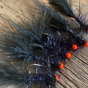 May include: Close-up of several black fishing flies with orange beads and silver hooks. The flies have dark, feathery plumes and are arranged on a wooden surface. The flies are designed for fly fishing.