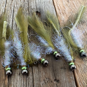 May include: A collection of six fishing flies. Each fly features a silver hook, a silver bead, and a green bead. The flies have olive green and iridescent blue-white synthetic fibers. The flies are arranged on a weathered wooden surface.