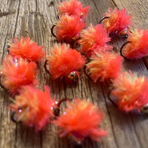 May include: Close-up of several bright orange and yellow fishing flies with dark hooks and silver beads. The flies are arranged on a weathered wooden surface, creating a textured background.