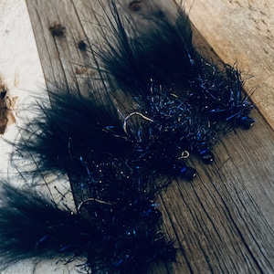May include: Close-up of three black fishing flies with iridescent blue accents. Each fly features a hook and is made with dark feathers and shiny, blue tinsel-like material. The flies are arranged on a weathered wooden surface.