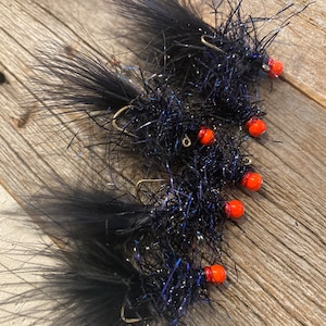 May include: Close-up of several black fishing flies with orange heads and gold hooks. The flies are made of fluffy, dark material with iridescent strands. They are arranged on a weathered wooden surface.