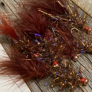 May include: Close-up of two handmade fishing flies. The flies feature reddish-brown feathers, iridescent tinsel, and copper-colored beads. The flies are designed for fly fishing and are arranged on a wooden surface.