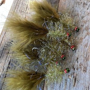 May include: Close-up of several handmade fishing flies. Each fly features a hook, olive-green feathers, and iridescent tinsel. The flies have small red and green beads near the hook eye. The flies are arranged on a weathered wooden surface.