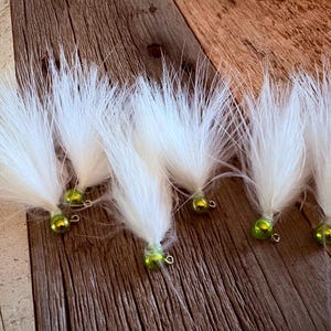 May include: A collection of six fishing lures, each featuring a bright lime green spherical jig head and a dense tuft of long, white synthetic fibres extending outward. Each lure includes a small metal eyelet attached to the jig head.