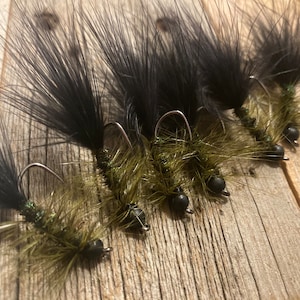 May include: Close-up of five fishing flies. Each fly features a black, feathery tail, a green body, and a black bead. The flies are arranged on a wooden surface, showcasing their intricate details and craftsmanship. Ideal for fly fishing enthusiasts.