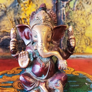 K&ouml;nnte beinhalten: Eine kleine, verzierte Ganesha-Statue in Gold und Rot mit einem nach oben gerollten R&uuml;ssel und einer Krone auf dem Kopf. Die Statue steht auf einem bunten, gemusterten Sockel.