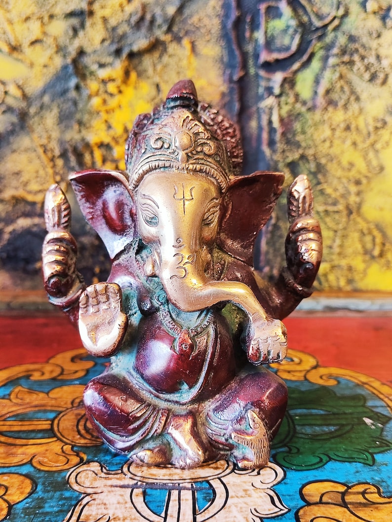 K&ouml;nnte beinhalten: Eine kleine, verzierte Ganesha-Statue in Gold und Rot mit einem nach oben gerollten R&uuml;ssel und einer Krone auf dem Kopf. Die Statue steht auf einem bunten, gemusterten Sockel.