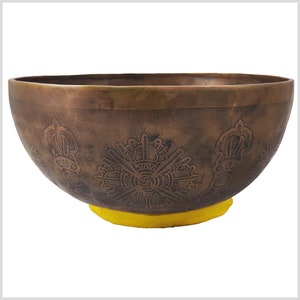 Pode incluir: Uma tigela de canto marrom com desenhos gravados intrincados, incluindo um mandala central e símbolos repetidos. A tigela tem uma base de borracha amarela.