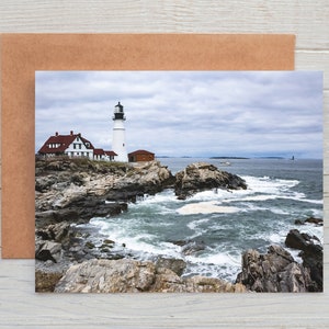 May include: A scenic photograph of a lighthouse on a rocky coastline under a cloudy sky. The white lighthouse stands tall next to a red-roofed building. The ocean waves crash against the rocks, creating a dramatic coastal scene.