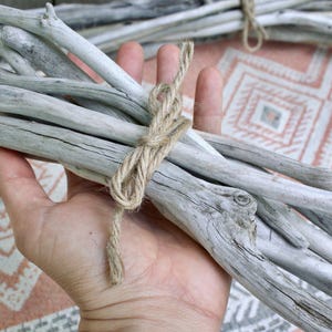May include: A bundle of weathered driftwood sticks tied together with twine. The gray-toned wood has a natural, textured appearance. The twine is a light brown color. The background is a patterned fabric in shades of pink, white, and gray.