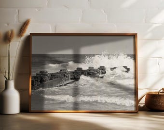 May include: Black and white framed print of ocean waves crashing against a stone breakwater. The photograph captures the power of the sea, with foamy white water and a dark, moody sky. The frame is a warm brown color.