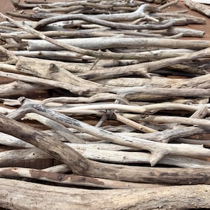 May include: A collection of weathered driftwood pieces in various sizes and shapes. The wood ranges in color from light beige to dark brown, with visible grain and texture. The pieces are arranged on a wooden surface, suitable for crafts or decor.