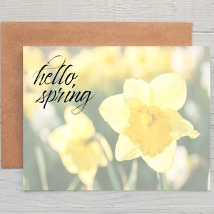 May include: A greeting card with a floral design featuring yellow daffodils and the words "hello, spring" in black script. The card is accompanied by a brown envelope, set against a light wood grain background.
