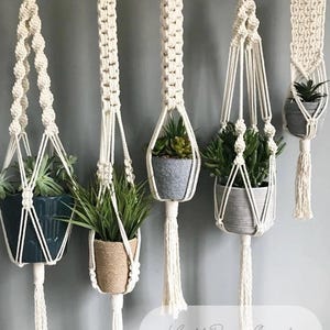 May include: Five off-white macrame plant hangers, each holding a different potted plant. The pots are various colors and textures, including blue, brown, and gray. The hangers are suspended from a gray wall, showcasing a bohemian decor style.