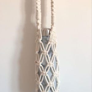May include: A light blue water bottle with a silver lid is held in a cream-colored macrame carrier. The carrier has a diamond pattern and two long handles for carrying. The background is a plain white wall.