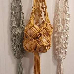 以下が含まれることがあります： セージグリーン、マスタードイエロー、オフホワイトのマクラメ吊り下げ式野菜バッグ3つ。黄色のバッグには玉ねぎが入っています。各バッグは上部に木製のリング、下部にタッセルが付いています。