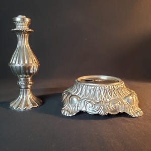May include: Two-piece silver-toned candlestick holder. The candlestick has a fluted design and a decorative base. The base is ornate with scrolled details. The pieces are separate, ready for assembly.
