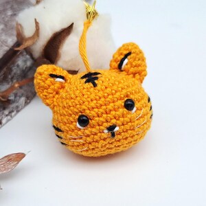 Può includere: Una testa di tigre arancione all'uncinetto con strisce nere e piccoli occhi neri. La testa di tigre ha un filo d'oro attaccato per appenderla. Lo sfondo include cotone e piante essiccate.