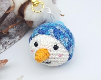 May include: A handmade crochet snowman keychain with a blue hat and an orange carrot nose. The snowman has black eyes and a small smile. The keychain has a gold-colored clasp.
