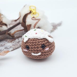 May include: A crocheted brown ornament with a smiling face and white frosting-like topping, accented with colorful sprinkles. It has a gold-colored keychain clasp. The ornament is set against a white background with cotton and wood accents.