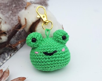 May include: A green crocheted frog keychain with a gold-colored clasp. The frog has black eyes and pink cheeks. The keychain is set against a white background with cotton and dried leaves.