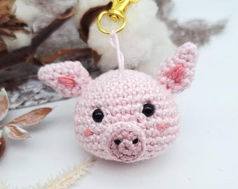 May include: A handmade, light pink crochet pig keychain with black eyes and pink accents on the ears and cheeks. The pig's snout is detailed, and it hangs from a gold-colored clasp. The background includes dried plants and cotton.