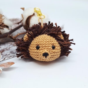 May include: A handmade crochet lion keychain. The lion has a tan face with black eyes and a black nose. The mane is made of brown yarn. A gold-colored clasp is attached to the top. The keychain is a cute accessory.