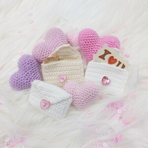 May include: A collection of crocheted heart-shaped pouches and envelopes in shades of pink, purple, and white. The pouches are decorated with buttons and some contain small gifts, such as rings and a card that says "I love you".